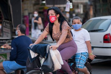 Rio de Janeiro, Brezilya - 3 Temmuz 2020: Kentin banliyölerindeki Coronavirus salgını sırasında insanlar maske takıyor. Oğlu sırtında bisiklet süren bir kadın..