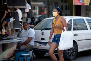 Rio de Janeiro, Brezilya - 3 Temmuz 2020: Kentin banliyölerindeki Coronavirus salgını sırasında insanlar maske takıyor. Alışveriş çantalı bir kadın geçerken bir erkek onu izliyor..
