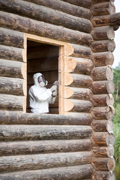 A worker in a white protective overalls and full face mask with anti-dust filters grinds a window opening in a freshly laid log house with a grinder.
