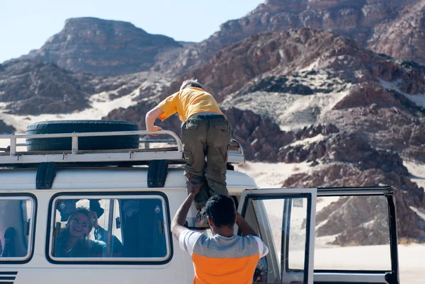 Jeep tour of Sinai in Egypt. The guide sits a boy in a yellow T-shirt on the roof of the car, where there is a spare wheel in the trunk. Mom's in the car laughing.