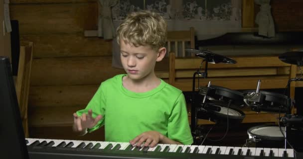 小男孩在电钢琴的黑色键上弹奏简单的旋律 一个金发男孩穿着绿色T恤 在白色和黑色的钢琴键盘上演奏 在木屋客厅的鼓包的背景 — 图库视频影像