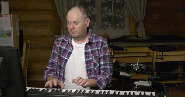 一个成熟的人在电钢琴上弹奏着节奏 在成年时学习 木屋里的客厅 在鼓包的背景下 一个人在一架电钢琴上拿起和弦 白色T恤和解开纽扣的格子花衬衫 秃头和胡茬 — 图库视频影像