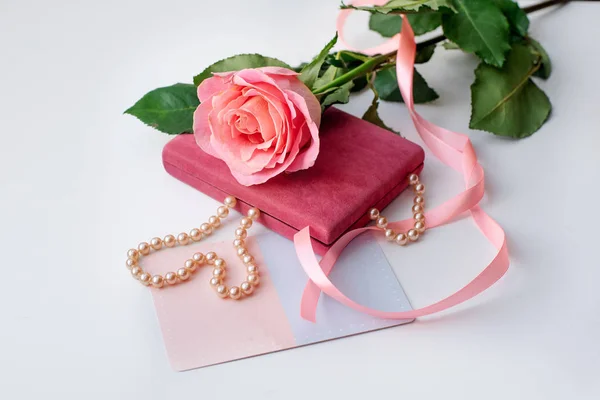 Pearl necklace on rose velvet box and pink one rose with gift card. Light background. Clouse-up.