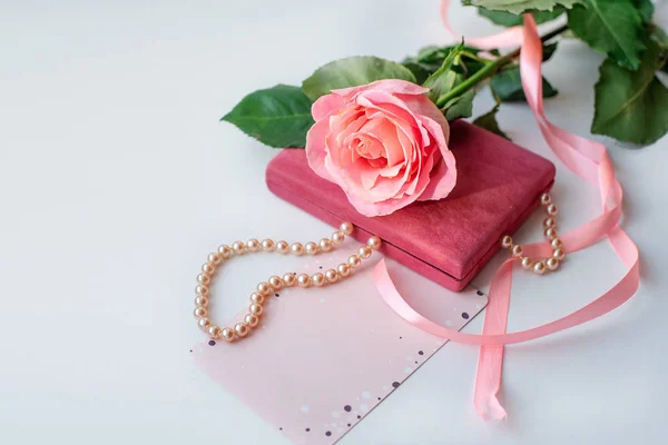 Pearl necklace on rose velvet box and pink one rose with gift card. Light background. Clouse-up.