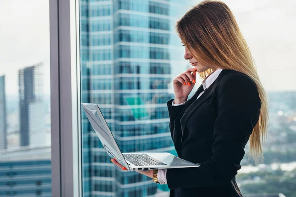 Pensativo CEO feminino em pé contra janela em seu escritório privado no moderno centro de negócios segurando um laptop leitura e pensamento — Fotografia de Stock