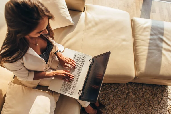 Vista superior de la mujer sentada en un sofá con una computadora portátil en su regazo escribiendo en el teclado en casa — Foto de Stock