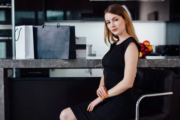 Portrait complet d'une femme élégante aux cheveux blonds portant une robe noire et des talons hauts assis sur une chaise de bar dans la cuisine — Photo