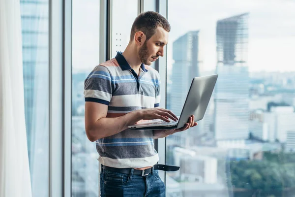 Homme d'affaires tenant un ordinateur portable travaillant debout dans le bureau près de la fenêtre avec vue sur la ville — Photo