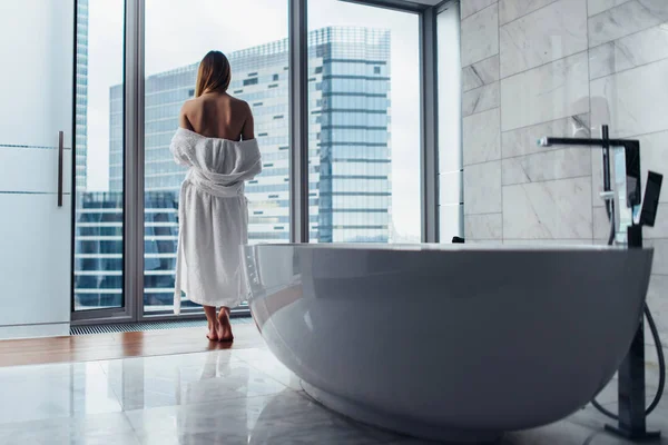 Вид ззаду молода жінка в білому халаті, що стоїть у ванній кімнаті з ванною на передньому плані — стокове фото