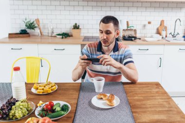 Cep telefonu kullaranarak mutfağında oturan adam.
