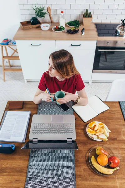 Femme tenant tasse assis à table avec ordinateur portable devant elle dans la cuisine . — Photo
