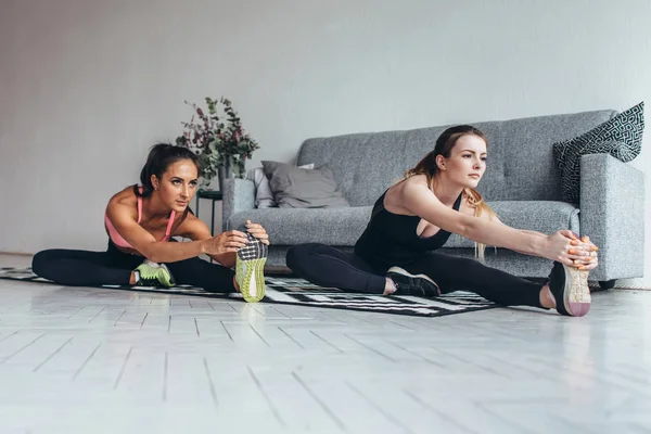 Ajustar a las mujeres haciendo estiramiento mientras están sentadas en el suelo en casa —  Fotos de Stock