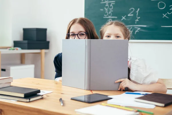 Écolières se cachant derrière le livre assis dans une salle de classe — Photo