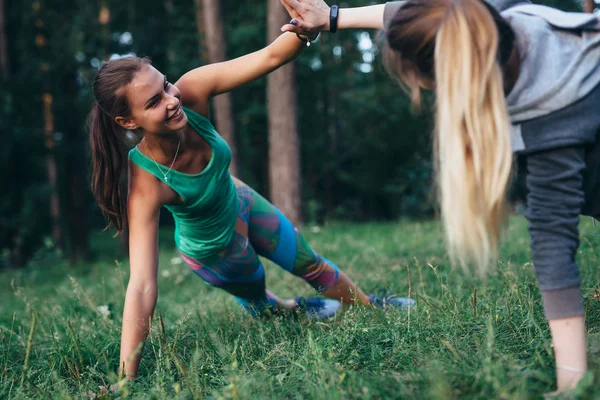 Duas jovens mulheres sorrindo trabalhando na grama no campo de saúde fazendo prancha lateral motivando uns aos outros segurando as mãos juntas — Fotografia de Stock