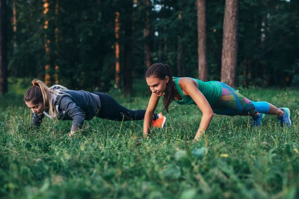 Duas amigas trabalhando juntas na floresta fazendo exercícios push-up na grama — Fotografia de Stock