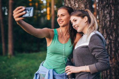 Selfie smartphone ile ormanda alarak ağacına yaslanmış spor giyim giymiş iki genç sportif kız arkadaşım