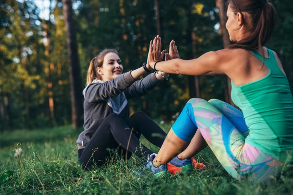 Twee positieve vriendinnen doen partner sit-up oefenen met high-five in zomer park — Stockfoto