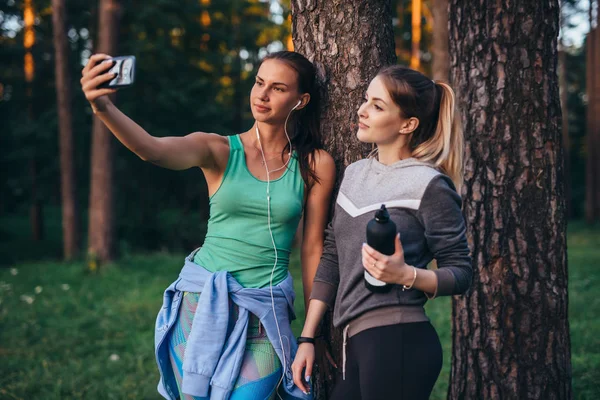 Dos jóvenes novias deportivas usando ropa deportiva apoyadas en el árbol tomando selfie con smartphone en el bosque —  Fotos de Stock