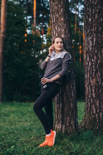 Retrato de una deportista bastante en forma usando un chándal apoyado en un árbol escuchando música en auriculares sosteniendo una botella de agua deportiva parada en el bosque — Foto de Stock