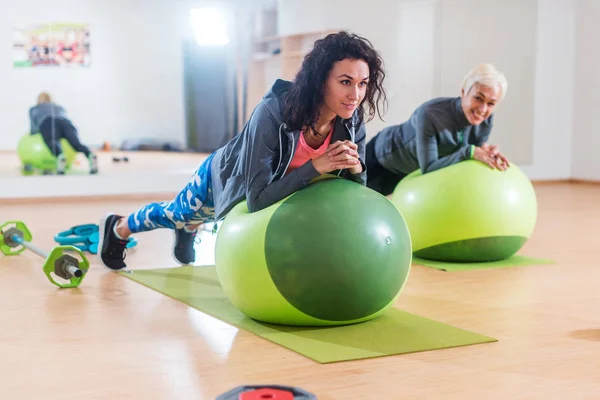 Dvě pozitivní ženy dělá prkno cvičení ležící na vyvážení míče v tělocvičně — Stock fotografie