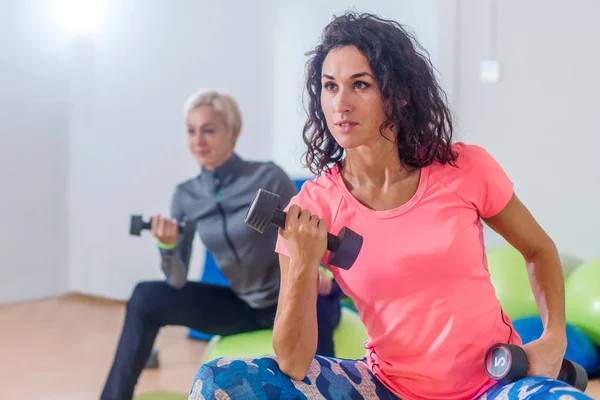 Sportliche schlanke Frauen, die an Fitness-Kursen teilnehmen und auf Physiobällen sitzend abwechselnd Bizeps-Curl mit Hanteln üben — Stockfoto