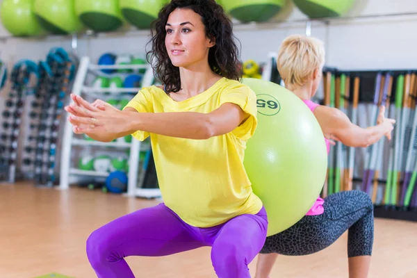 Zwei junge Frauen, die Kniebeuge-Übungen machen, mit einem schweizerischen Ball dazwischen. Sportlerinnen trainieren im Fitnessstudio. — Stockfoto