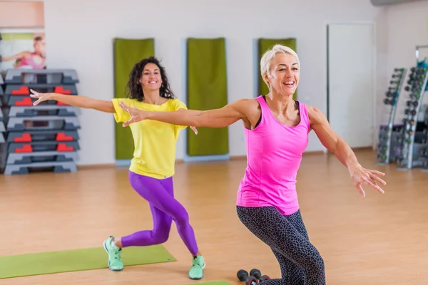 Duas mulheres atléticas sorridentes fazendo exercícios de dança aeróbica segurando os braços para o lado dentro de casa no centro de fitness . — Fotografia de Stock