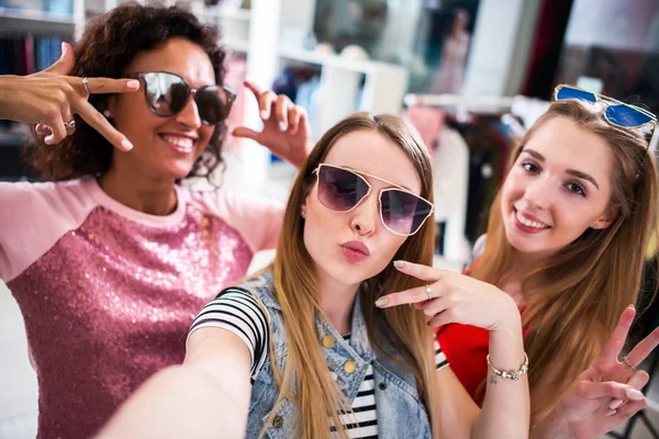 Meninas bonitas usando óculos de sol brincando com selfie mostrando gestos de língua e chifre na loja de roupas — Fotografia de Stock