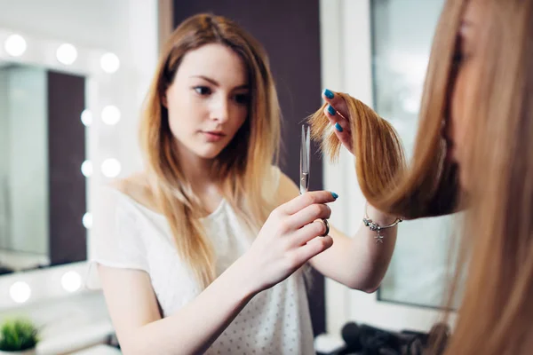 Young pretty hairdresser wearing casual clothes holding strand of long hair between fingers trimming the ends in beauty studio — Stock Photo, Image