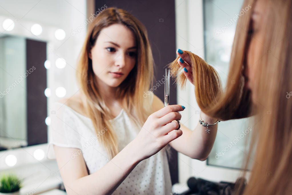 Young pretty hairdresser wearing casual clothes holding strand of long hair between fingers trimming the ends in beauty studio