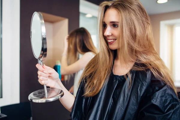 Joven cliente femenina con capa sosteniendo un espejo de maquillaje mirando y sonriendo en el salón de belleza satisfecho con el maquillaje y corte de pelo — Foto de Stock