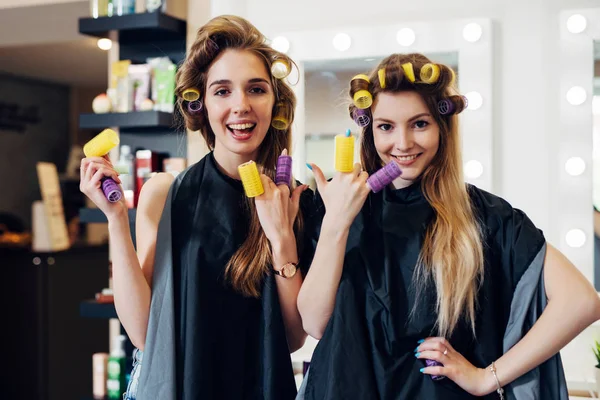 Två flickor curling håret med papiljotter bär cape ha kul skrattande att sätta rullarna på fingrarna i frisörsalong — Stockfoto
