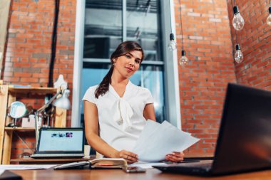 Genç kadın gazeteci holding kağıt yaratıcı ofis masasında otururken dizüstü kullanarak yeni bir makale hazırlama