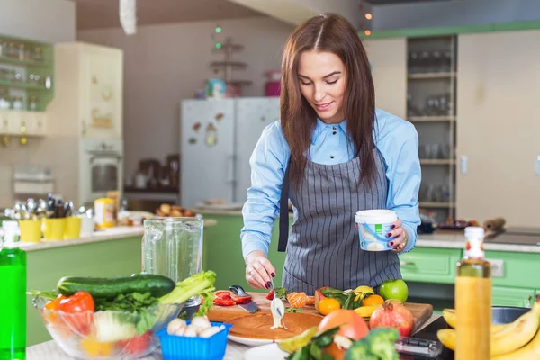 Glad ung kvinna att göra tårta smetas ut ett lager med grädde stående i köket — Stockfoto