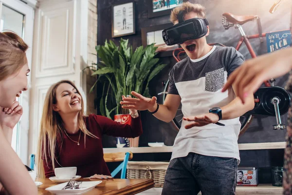 Chico joven probando auriculares de realidad virtual gritando jugando juego de miedo mientras sus alegres amigas guapas se ríen de él sentadas en un café — Foto de Stock