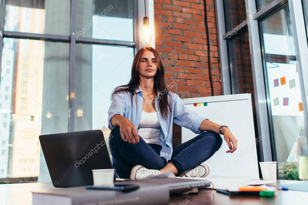 Female student sitting in lotus pose on table in her room meditating relaxing after studying and preparing for exam