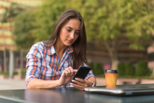Ελκυστική νεαρή γυναίκα που διαβάζετε ένα μήνυμα κειμένου στο κινητό της. Κορίτσι που κάθεται σε εξωτερικούς χώρους χρησιμοποιώντας το smartphone — Φωτογραφία Αρχείου