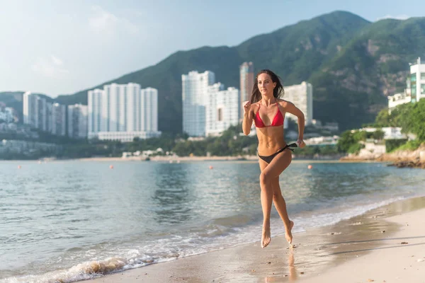 Vista posterior de ajuste chica delgada corriendo descalzo en la orilla del mar con bikini. Mujer joven haciendo ejercicio cardio playa iluminada al sol y montañas de la ciudad en el fondo . —  Fotos de Stock