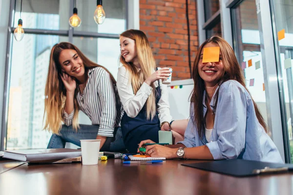 Dos chicas riéndose de su amiga con una nota pegajosa en la cara. Grupo de alumnas relajándose divirtiéndose en el aula durante un descanso — Foto de Stock