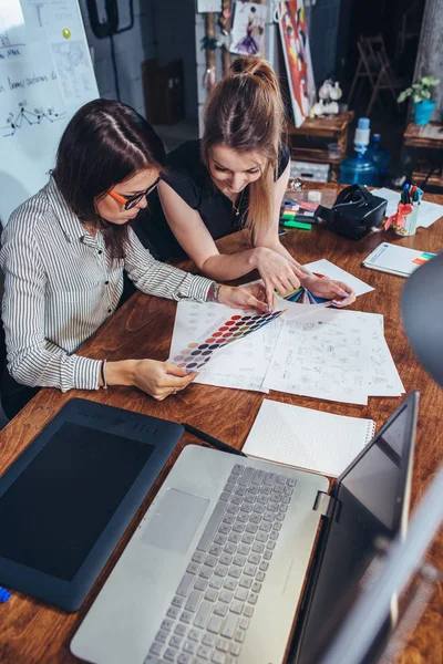 Unga kvinnliga grafiska designers sitter vid skrivbord med laptop, färgkartor och några skisser som diskuterar det nya projektet att välja ett koncept — Stockfoto