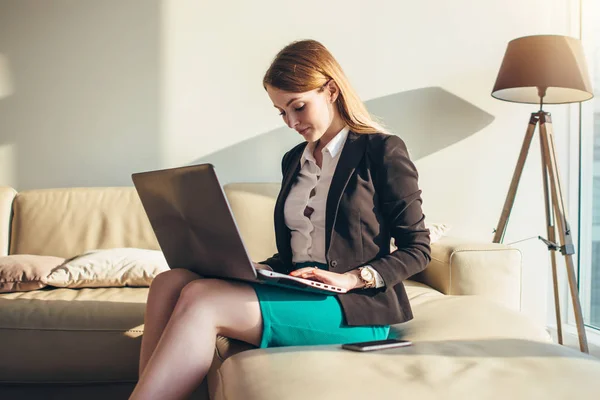 Женщина сидит на диване с ноутбуком на коленях и печатает дома на клавиатуре — стоковое фото
