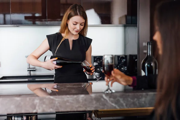 Young women wearing elegant black dress holding a bottle of red wine and a glass standing at kitchen bar looking at her female friend — Stock Photo, Image