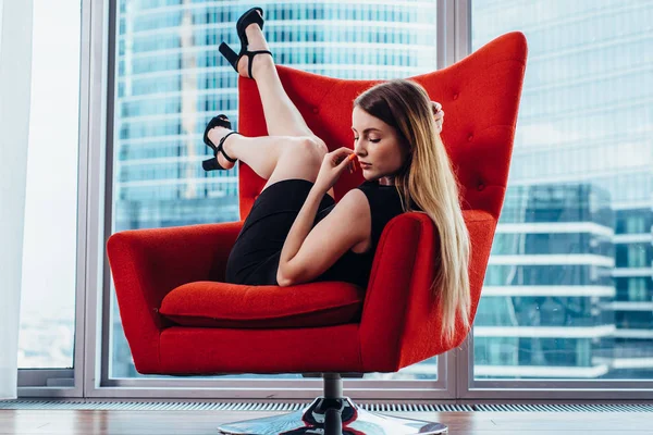 Modelo femenino sexy con pelo claro posando en un elegante sillón rojo contra una ventana panorámica con vista a rascacielos —  Fotos de Stock