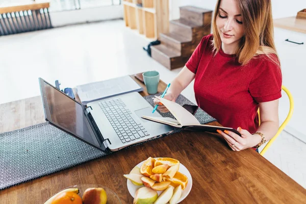 Mulher escrevendo e tomando notas, usando seu laptop na cozinha. Trabalhando em casa conceito — Fotografia de Stock