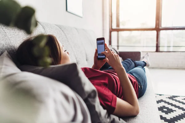 Junge Frau benutzt Smartphone, während sie zu Hause auf der Couch liegt — Stockfoto