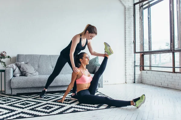 Подходящая женщина делает упражнения на растяжку с помощью подруги, держащей ногу дома — стоковое фото