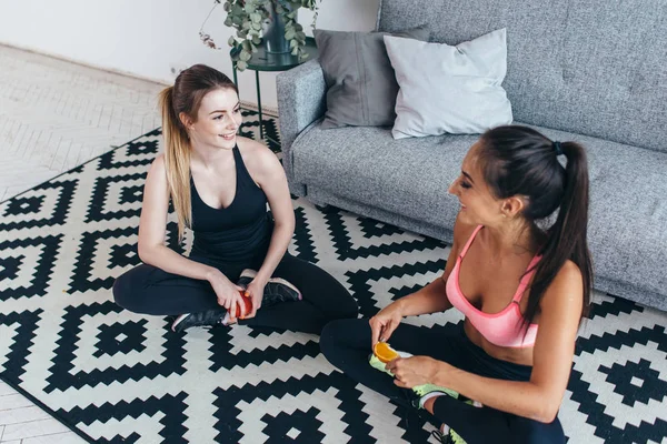 Sonrientes mujeres en forma usando ropa deportiva sentadas en el suelo comiendo frutas y hablando — Foto de Stock