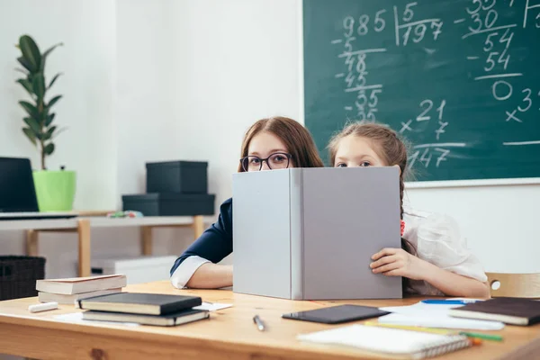Bir sınıfta oturup kitap arkasına saklanmış kız öğrenciler — Stok fotoğraf