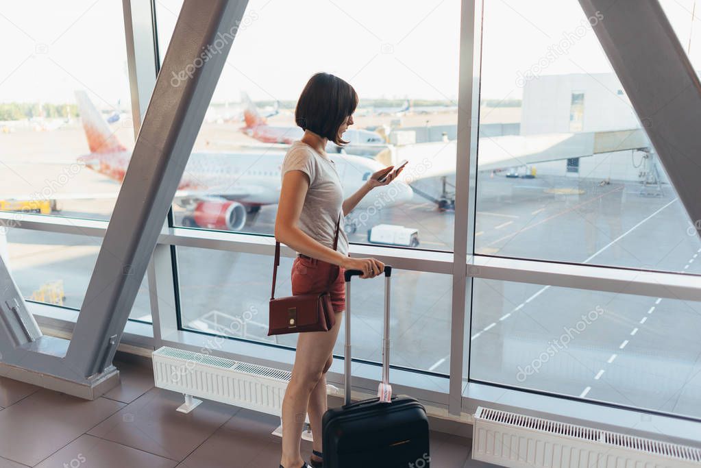 Young casual female traveler in airport, holding smart phone near gate windows at planes on runway.