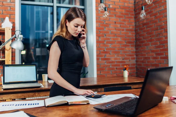 かなり若い女性の電話を数える作業デスクで事務所に立っているを計算機を使用しての話 — ストック写真